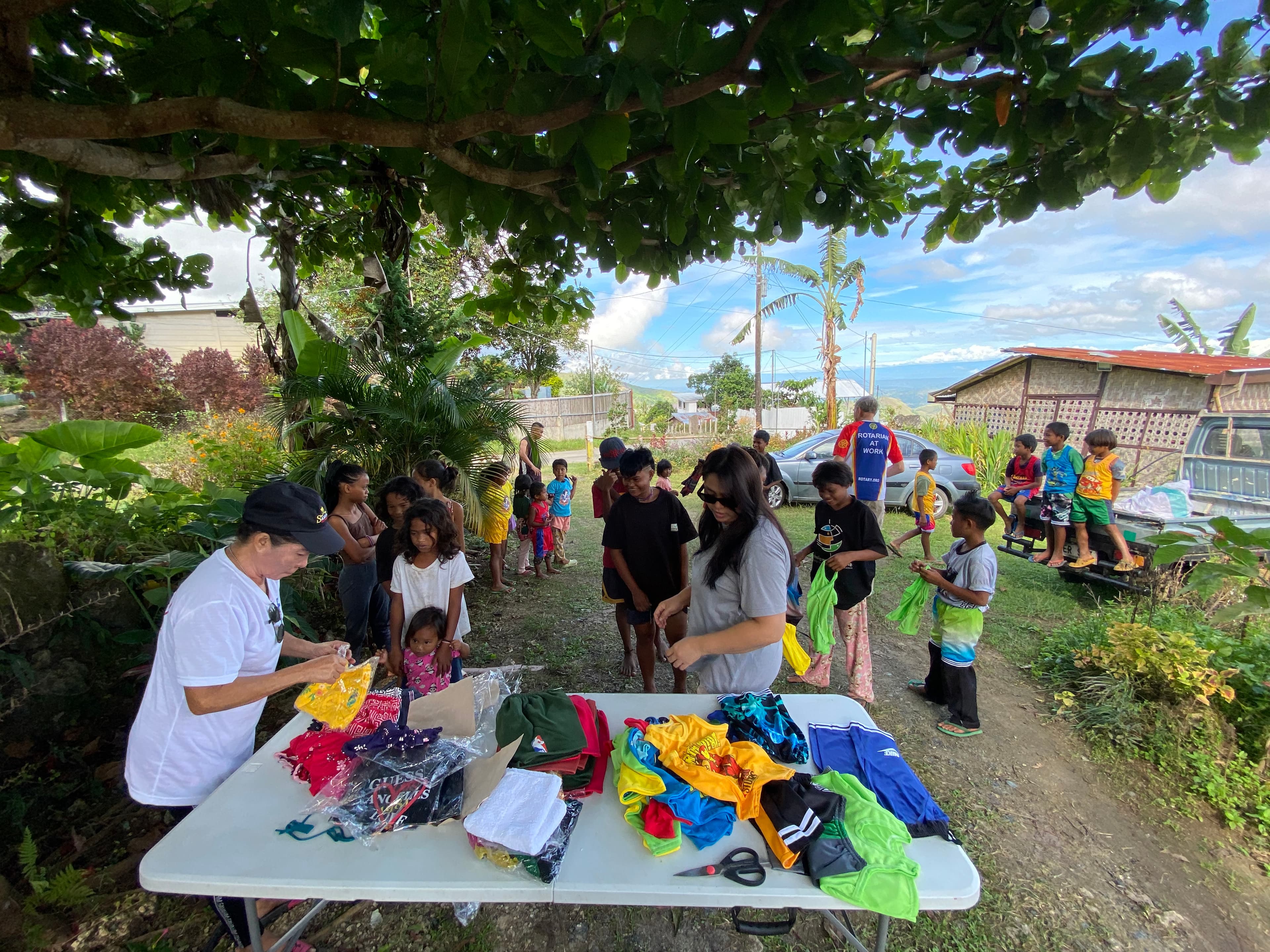 Distribution day under a tree — families coming up to a table of folded clothes